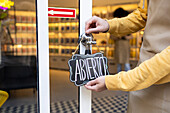 A person in a yellow jacket putting up an 'Abierto' sign, signaling the opening of a business, with a reflective glass door backdrop.