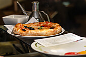 A deliciously baked pizza garnished with black olives and fresh basil oil, served on a ceramic plate alongside condiments, creating a tempting culinary presentation