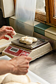 A person's hands are shown weighing a ball of fresh pizza dough on a red digital scale in a rustic kitchen setting, capturing a step in the pizza-making process