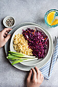 Ein nahrhaftes Mittagessen mit einem leuchtenden Salat aus rohem Rotkohl und Karotten sowie lockerem gekochtem Bulgur, serviert auf einem Keramikteller mit frischen grünen Zwiebeln und einem Zitruswasser an der Seite