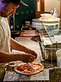 A professional chef carefully adds cheese to a freshly sauced pizza base in a well-equipped commercial kitchen, highlighting the art of pizza making