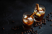 From above two glasses of chilled coffee adorned with cinnamon sticks and floating ice cubes, set against a backdrop of scattered coffee beans on a dark surface.