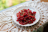 A bowl of ripe redcurrant berries on a beautifully patterned white lace tablecloth, sitting beside a window with natural light