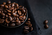 From above close-up view of roasted coffee beans in a dark bowl on a textured black surface, highlighting the rich textures and colors.