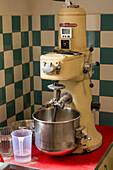 An aged dough mixer stands on a red tabletop, backed by green and cream tiled walls, equipped for culinary use with simple attachments and a mixing bowl