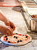 A close-up image showing a chef's hands topping a pizza with mozzarella cheese and black olives The chef skillfully works on a marble counter with tomato sauce and ingredients around