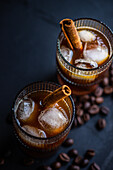 From above, two glasses of chilled coffee adorned with cinnamon sticks and floating ice cubes, set against a backdrop of scattered coffee beans on a dark surface.