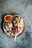 A rustic ceramic bowl holds whole garlic bulbs next to a spoon filled with vibrant red paprika, set against a textured grey background