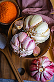 A rustic culinary scene with garlic in a wooden bowl next to a bright orange spice bowl, all placed on a wooden table with a soft linen background