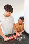Two siblings enjoy baking together in the kitchen, with one using a rolling pin on dough while the other laughs The scene captures the joy and bonding of cooking as a fun, shared activity
