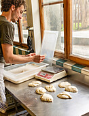 A baker meticulously weighs portions of pizza dough on a digital scale, ensuring consistent quality, amidst the surroundings of a natural lit, rustic kitchen