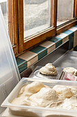 The image captures fresh pizza dough balls proofing on scales and resting in a plastic container by a sunny window, showcasing the initial steps of pizza preparation in a rustic kitchen setting