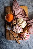 A rustic culinary scene featuring whole garlic bulbs arranged artistically on a wooden board, accompanied by a bowl of vibrant orange spices and a pinch of salt, all set against a textured grey backdrop with a napkin