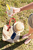 A person holding a glass of lemonade with a lemon slice, next to a straw hat, sunglasses, and phone on a bag, symbolizing a relaxed summer day