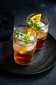 Two glasses of a refreshing summer cocktail featuring cherry liqueur mixed with tonic, beautifully garnished with orange slices, ice cubes, and fresh mint leaves on a dark, textured surface