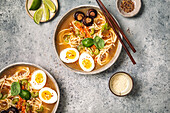 Ramen bowls with noodles, eggs, mushrooms, carrots and sesame seeds on grey background