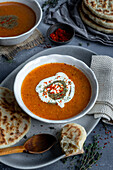 Tarhana soup topped with yogurt, dried mint and red pepper flakes in a white bowl, some flat bread and a wooden spoon accompany.