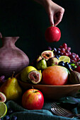 Seasonal Autumn Fruit Bowl with Apples, Pears, Mangoes, Figs, Limes, and Grapes. Female hand holding apple.