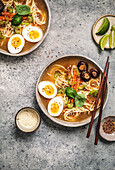 Ramen bowls with noodles, eggs, mushrooms, carrots and sesame seeds on grey background