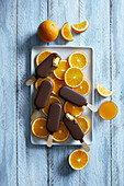 Chocolate orange popsicle ice cream arranged on a white plate with freshly cut orange slices on a blue wooden background