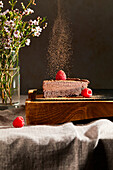 Dusted cocoa powder falling on a slice of Lisbon Chocolate Cake with raspberries as a garnish on a table with flowers in the background.