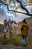 Rückenansicht einer jungen Frau mit gelber Jacke, die auf die mittelalterliche historische Burg Eltz in einer Herbstlandschaft mit Bäumen bei Sonnenaufgang blickt, Wierschem, Rheinland-Pfalz, Deutschland, Europa