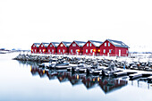 Reihe roter traditioneller Holzhütten über einem kleinen Hafen in der verschneiten Landschaft, Leknes, Lofoten, Norwegen, Skandinavien, Europa