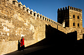 Frau in rotem Mantel vor der historischen Muralla de la Macarena in Sevilla. Sonnenbeschienene alte Mauer mit Schatten.