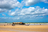 Touristen erkunden den Goldstrand in Arromanches, Normandie, Frankreich, mit den Überresten des Hafens von Mulberry B im Hintergrund.