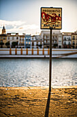 A graffiti-covered 'Prohibido Bañarse' sign on the riverbank in Sevilla, España, with colorful buildings in the background.