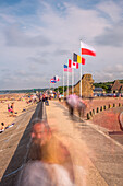 Langzeitaufnahme der Promenade am Omaha Beach in der Normandie, Frankreich, mit verschiedenen Nationalflaggen und Strandbesuchern.