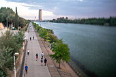 People strolling and cycling along a riverside walkway with Torre Sevilla in the background during dusk. A serene urban scene in Sevilla.