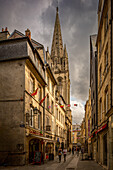 Malerische Straßenansicht von Caen mit der hoch aufragenden Kirche Saint Sauveur in der Normandie, Frankreich. Malerische Stadtatmosphäre mit Fußgängern und lokalen Geschäften.