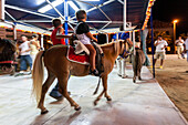 Kinder genießen eine Ponyfahrt auf einem Nachtkarussell in Sanlucar de Barrameda, Andalusien. Spaß und Aufregung auf einem lokalen Jahrmarkt.