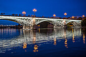 Nächtlicher Blick auf die beleuchtete Triana-Brücke über den Fluss Guadalquivir in Sevilla, Spanien. Die Reflexionen auf dem Wasser und die Lichter der Stadt schaffen eine heitere Atmosphäre.