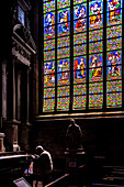 A man prays by the stained glass windows in Collegiale Saint Aubin, Guerande, France. The colorful windows depict religious scenes.