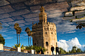 Atemberaubender Blick auf den Torre del Oro, der sich in einer Pfütze nach einem Regen in Sevilla, Spanien, spiegelt. Der klare blaue Himmel hebt die leuchtenden Farben und historischen Details hervor.