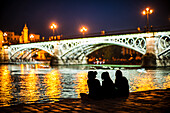 Eine Gruppe von Freunden genießt einen Abend am Flussufer in der Nähe der wunderschön beleuchteten Triana-Brücke in Sevilla, Spanien. Aufgenommen mit einem Leica Noctilux Objektiv.