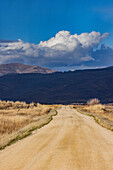 Empty dirt road leading to foothills under stormy skies