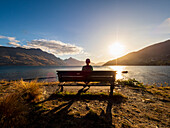 Rückansicht einer Frau, die bei Sonnenuntergang auf einer Bank mit Blick auf den Lake Wakatipu sitzt