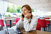 Frau mit Smartphone, Kopfhörern und Laptop an einem Cafétisch