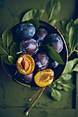 Whole and halved plums with leaves in a bowl