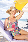 Woman in floral swimming costume and straw hat on beach under parasol, drink