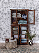 Antique display cabinet and wicker basket in front of floral patterned wallpaper