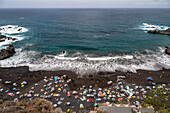 Aerial view of Playa del Bollullo in Tenerife with beachgoers, black sand, and crashing ocean waves. Located in La Orotava, Canary Islands, Spain.