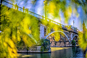 Scenic view of Triana Bridge framed by lush greenery along the riverbank in Seville, Andalucia, Spain. Captured with a Leica Noctilux lens, highlighting architecture and nature's beauty.