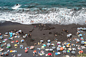 Aerial view of Playa del Bollullo in Tenerife with beachgoers, black sand, and crashing ocean waves. Located in La Orotava, Canary Islands, Spain.