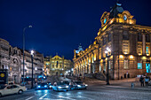 Evening scene of Rua das Flores street featuring Sao Bento railway station and Sao Antonio dos Congregados church in Porto, Portugal, with illuminated buildings and traffic.