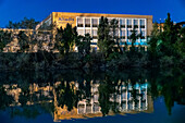 Seville, Spain, Jan 12 2017, A nighttime view of the disused tobacco factory in Sevilla, Spain, with its reflection cast in the Guadalquivir River, surrounded by trees.