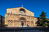 Tourists admire the stunning facade of Santo Domingo Church in Soria, featuring intricate Romanesque design elements against a clear blue sky.